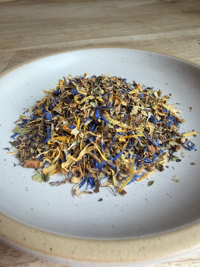 A close-up top-down view of loose dried herbal tea or flower blend piled in the center of a white ceramic bowl, with visible blue, yellow, brown, and green botanical pieces.