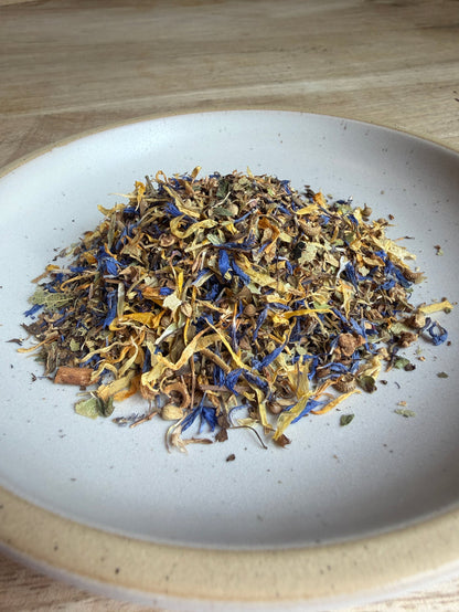 A close-up top-down view of loose dried herbal tea or flower blend piled in the center of a white ceramic bowl, with visible blue, yellow, brown, and green botanical pieces.