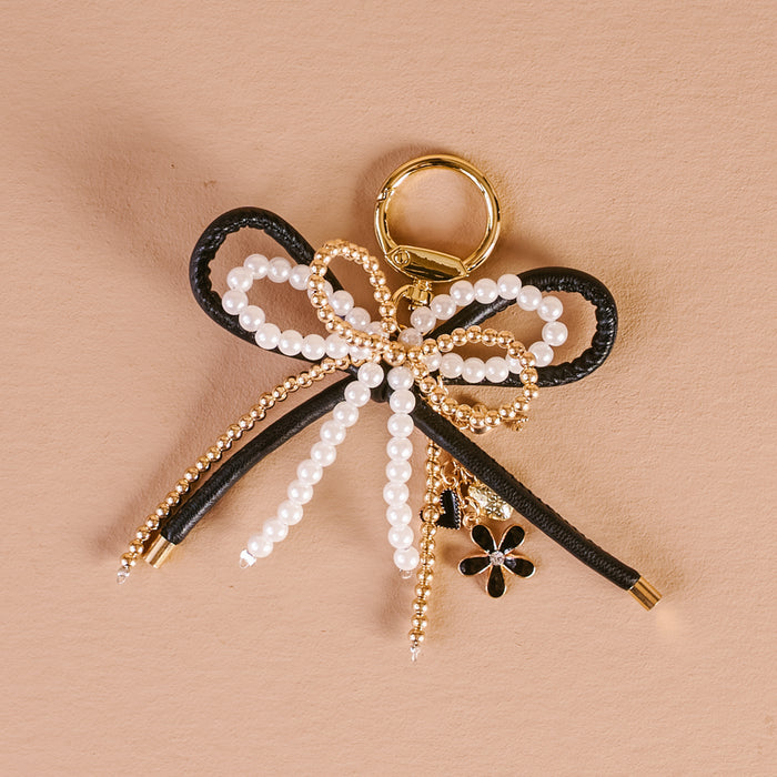 The Darling Effect’s Luxe Glam Layered Bows Bag Charm features a gold ring, black cord bow, gold beads, white pearls, and a black and gold flower charm—an elegant accessory on a textured beige background.