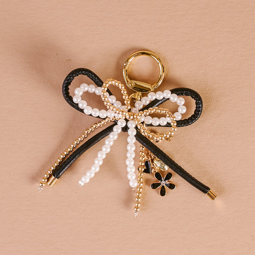 The Darling Effect’s Luxe Glam Layered Bows Bag Charm features a gold ring, black cord bow, gold beads, white pearls, and a black and gold flower charm—an elegant accessory on a textured beige background.