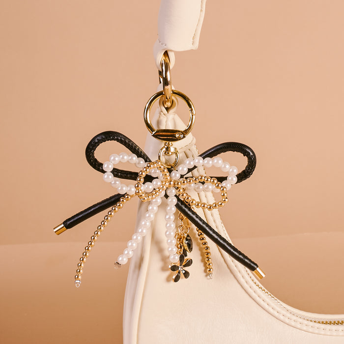 The Darling Effect's Luxe Glam Layered Bows Bag Charm in black and white pearls with gold accents hangs from a cream strap, adding a chic statement to your handbag.