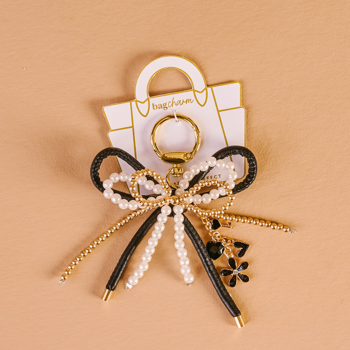 The Darling Effect’s Luxe Glam Layered Bows Bag Charm features black and gold loops, pearl beads, a black flower charm, and a gold chain, presented on a handbag-shaped card with a beige background.