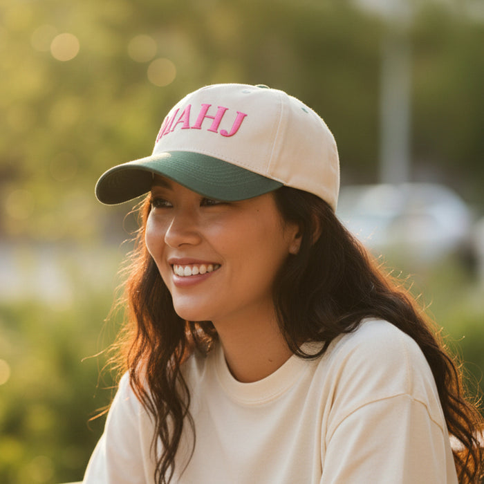 A smiling woman wears The Darling Effect’s Mahj Classic Canvas Hat—cream-colored canvas with pink “MAHJ” embroidery and a green brim—for effortless, everyday style.