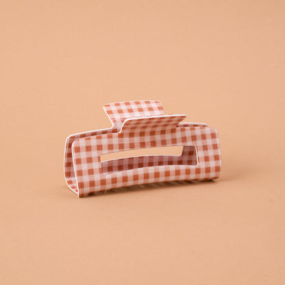 The Maple Gingham Claw Clip by The Darling Effect is a stylish rectangular hair accessory with rounded edges, featuring a charming pink and white gingham checkered pattern and displayed on a plain light brown background.