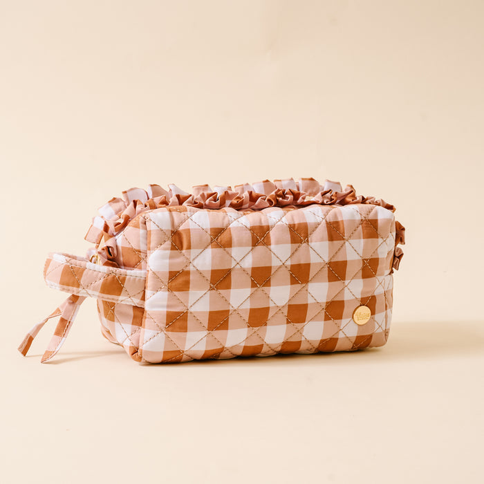 Quilted cosmetic pouch with a brown and white checkered pattern, ruffled top edge, side handle, and a small round gold emblem, featuring a wipeable interior for easy cleaning—beautifully placed on a beige background.