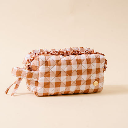 Quilted cosmetic pouch with a brown and white checkered pattern, ruffled top edge, side handle, and a small round gold emblem, featuring a wipeable interior for easy cleaning—beautifully placed on a beige background.