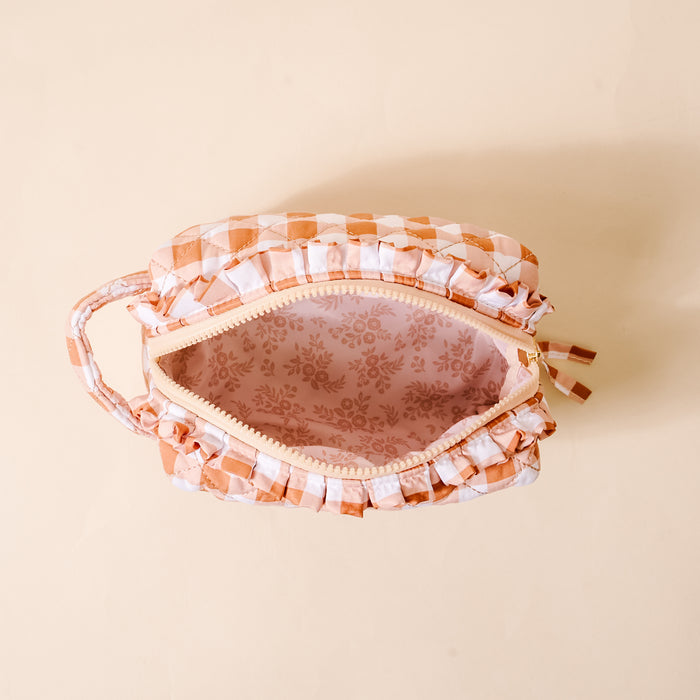 A top view of The Darling Effect Maple Gingham Essentials Quilted Cosmetic Pouch showcases its open, peach and white checkered design with a handle. The makeup bag reveals a pink floral-patterned, wipeable interior lining, set against a light beige background.