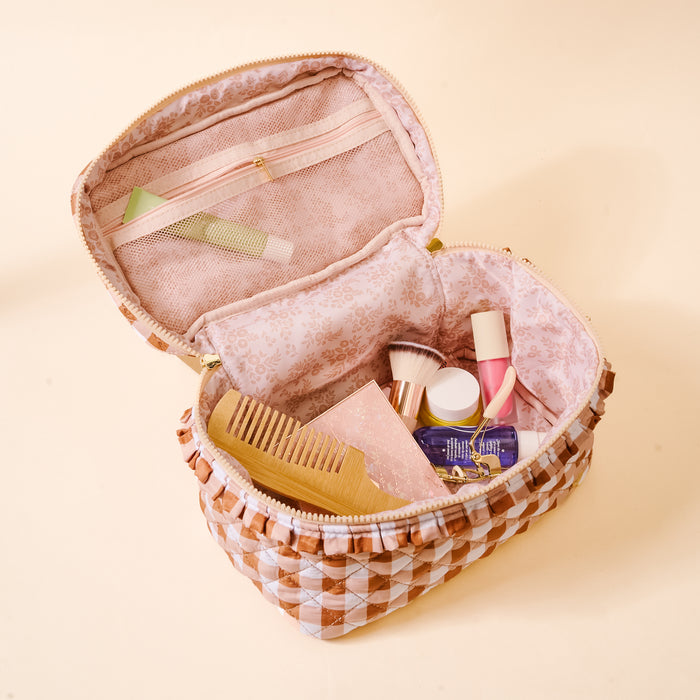The Maple Gingham Glam & Go Quilted Cosmetic Bag by The Darling Effect is open, revealing a wooden comb, makeup brushes, skincare bottles, and a pink box inside. This pink and white checkered bag features a floral lining and a small zippered pocket, making it the perfect travel organizer for all your beauty essentials.