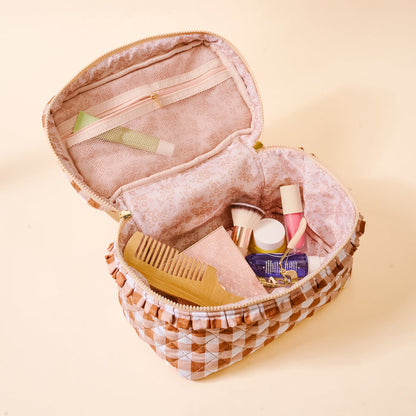 The Maple Gingham Glam & Go Quilted Cosmetic Bag by The Darling Effect is open, revealing a wooden comb, makeup brushes, skincare bottles, and a pink box inside. This pink and white checkered bag features a floral lining and a small zippered pocket, making it the perfect travel organizer for all your beauty essentials.