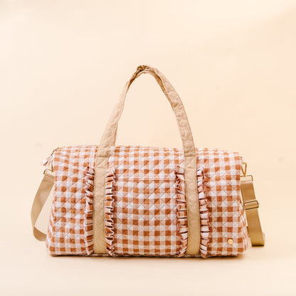 The Darling Effect’s Maple Gingham On-the-Go Quilted Weekender Bag is a spacious travel bag showcasing a beige and white gingham pattern with charming ruffle accents on the front, two beige handles, and a beige adjustable shoulder strap, displayed on a light beige background.