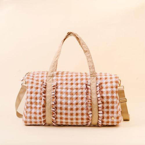 The Darling Effect’s Maple Gingham On-the-Go Quilted Weekender Bag is a spacious travel bag showcasing a beige and white gingham pattern with charming ruffle accents on the front, two beige handles, and a beige adjustable shoulder strap, displayed on a light beige background.
