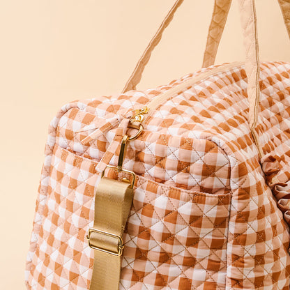 Introducing the Maple Gingham On-the-Go Quilted Weekender Bag by The Darling Effect, showcasing a spacious design with a tan and white gingham pattern, gold zipper accents, and an adjustable beige strap—beautifully photographed against a light beige background.
