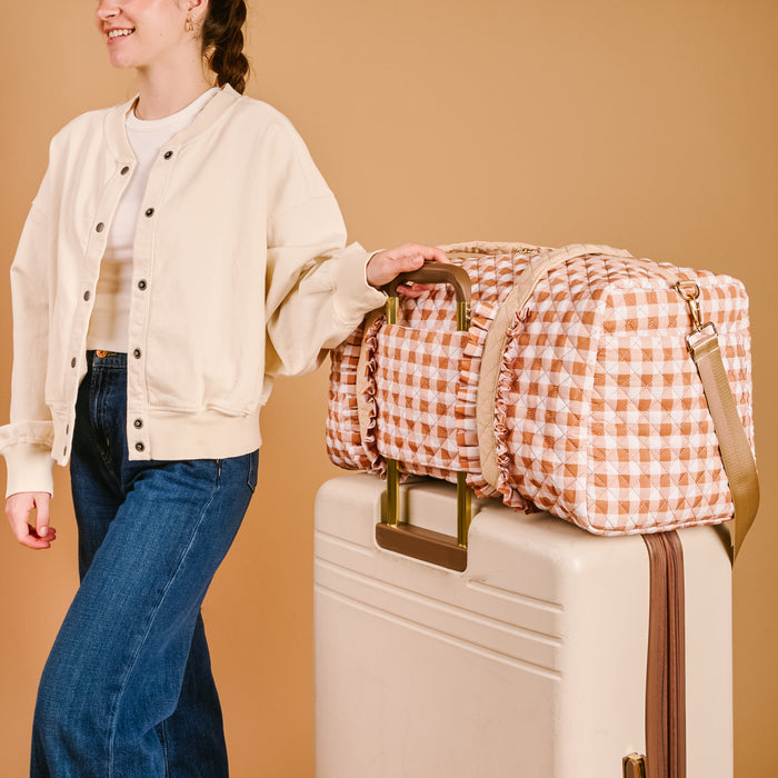 A person in a cream jacket and jeans stands next to a white suitcase topped with The Darling Effect’s Maple Gingham On-the-Go Quilted Weekender Bag, set against a beige background.