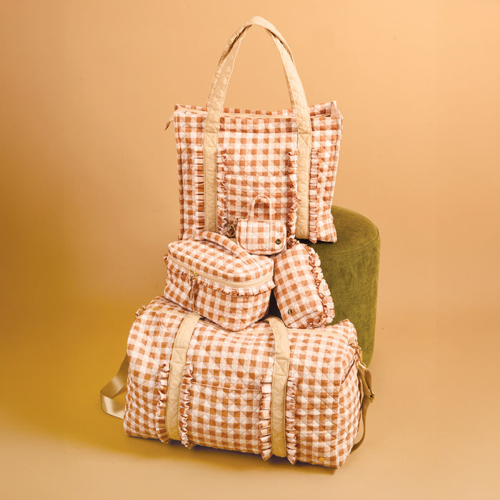 Five peach and white gingham-patterned bags, including The Darling Effect’s Maple Gingham Glam & Go Quilted Cosmetic Bag, a tote, duffel, travel organizer, and a small pouch, are arranged against a beige background beside a green round ottoman.