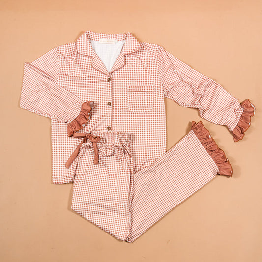 Experience cozy comfort and charming style with the Maple Gingham Sweet Dreams Pajama Pants Set from The Darling Effect. This pink and white checkered set features a long-sleeve button-up top paired with matching pants, both highlighted by playful ruffled cuffs for a feminine finish. The pants include a comfortable drawstring waist, making this set perfect for relaxing nights in or as an adorable gift option. Displayed on a beige background.