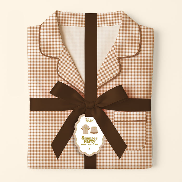 Folded brown and white Maple Gingham Sweet Dreams Pajama Shorts Set by The Darling Effect, tied with a dark brown ribbon and featuring a gift tag that reads “Slumber Party Pajama Set S,” displayed against a cream background—a perfect pajama gift set for cozy nights in comfortable pajamas.