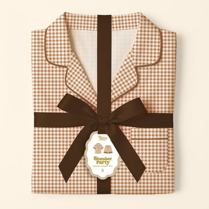 Folded brown and white Maple Gingham Sweet Dreams Pajama Shorts Set by The Darling Effect, tied with a dark brown ribbon and featuring a gift tag that reads “Slumber Party Pajama Set S,” displayed against a cream background—a perfect pajama gift set for cozy nights in comfortable pajamas.