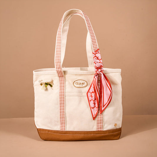 The Maple Gingham The Darling Canvas Tote Bag by The Darling Effect features a cream canvas body with a roomy interior, accented by a brown base, red plaid handles, a red patterned scarf tied to one handle, and a small decorative pin. It is pictured against a plain light brown background.