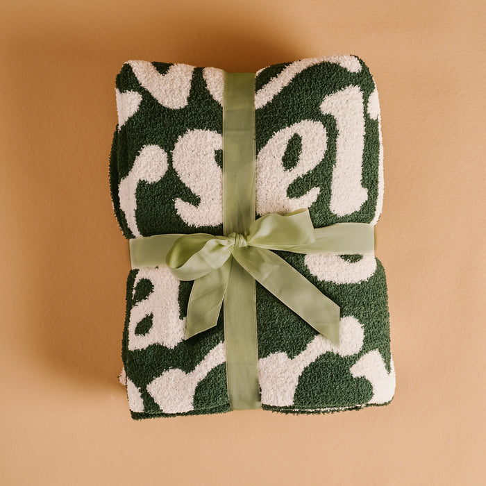 The Darling Effect's Merry Little Christmas Cozy Dreams Luxe Blanket, in a fluffy green and white pattern, is made of ultra-soft microfiber and tied with a light green ribbon on a tan background.