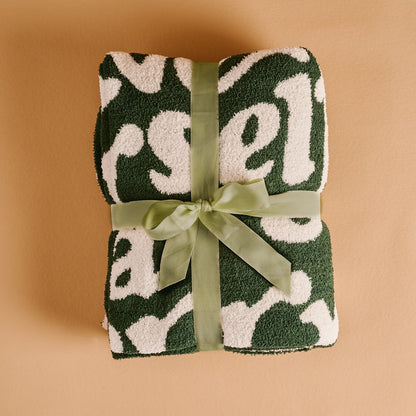 The Darling Effect's Merry Little Christmas Cozy Dreams Luxe Blanket, in a fluffy green and white pattern, is made of ultra-soft microfiber and tied with a light green ribbon on a tan background.