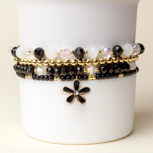 The Midnight Glam Tumbler Bracelet Stack by the-darling-effect features black, white, and gold beads with a gold flower charm, displayed on a white stand—perfect as accessories for your favorite 40 oz tumbler.