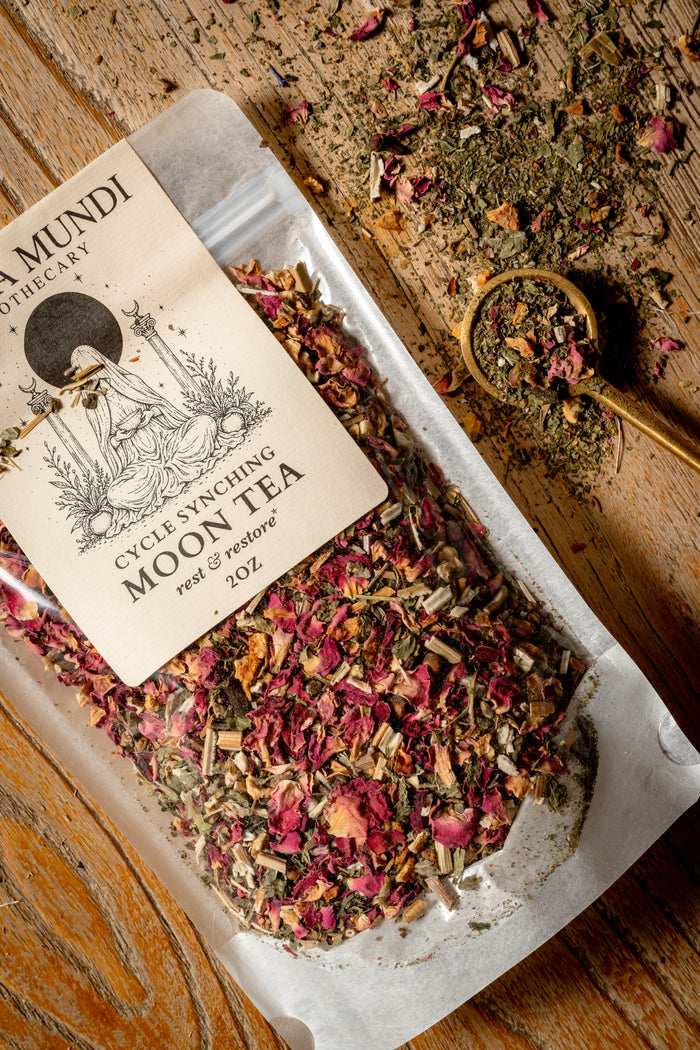 A 2oz stand-up pouch of Amundi Botanicals Cycle Syncing Moon Tea, filled with dried herbs and pink flower petals, displayed on a rustic wooden surface next to a wooden spoon filled with the tea.