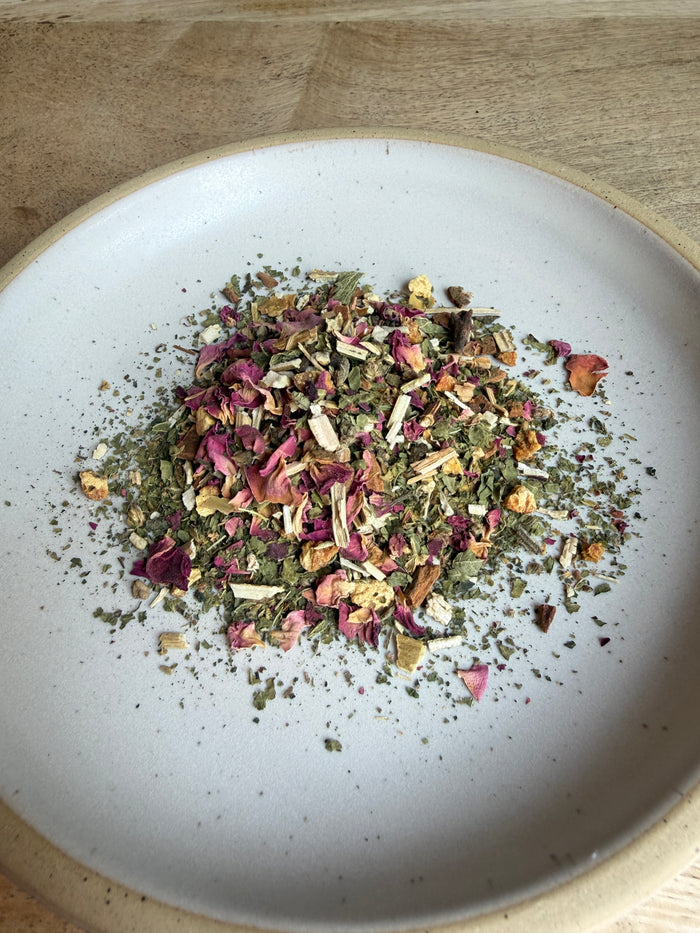 A close-up top-down view of loose-leaf herbal tea or dried botanicals piled in the center of a white ceramic plate with a beige rim, set on a light wood surface.