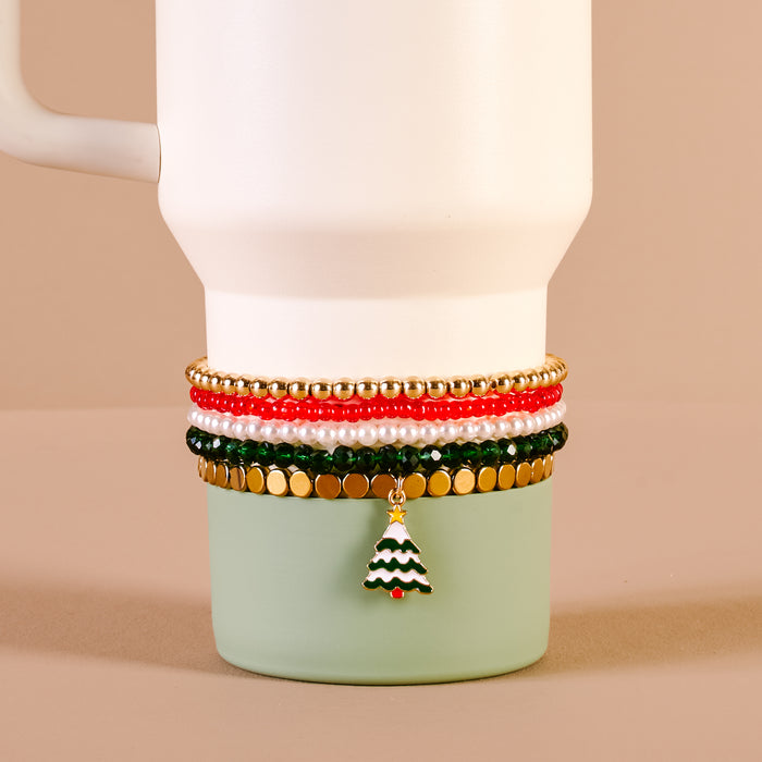 The Darling Effect’s O Christmas Tree Tumbler Bracelet Stack in gold, red, green, and white—featuring a Christmas tree charm—is styled on a white mug with green base against a neutral beige background. Perfect for pairing with tumbler accessories.