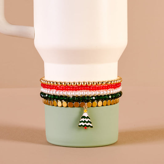 The Darling Effect’s O Christmas Tree Tumbler Bracelet Stack in gold, red, green, and white—featuring a Christmas tree charm—is styled on a white mug with green base against a neutral beige background. Perfect for pairing with tumbler accessories.