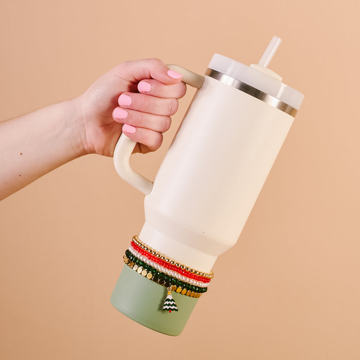 A hand with pink nails holds a 40 oz cream tumbler with a green base and straw, featuring The Darling Effect’s O Christmas Tree Tumbler Bracelet Stack wrapped around the bottom for playful accessorizing.