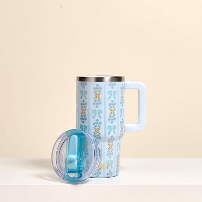The Oh So Fancy Blue 20 oz Lifestyle Flip Straw Tumbler by the-darling-effect, featuring a tall, floral-patterned design and large handle, stands on a white surface with its clear blue flip straw lid in front. The background is beige.