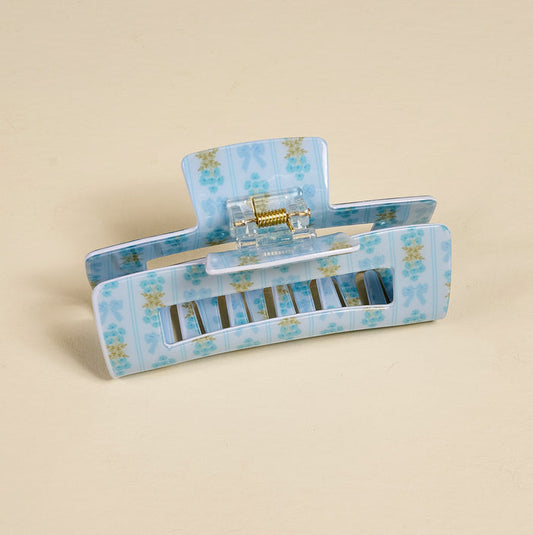 The Darling Effect’s Oh So Fancy Blue Claw Clip, a blue rectangular accessory with a floral print, gold-colored springs, and interlocking teeth for strong hold, is shown on a plain beige background.