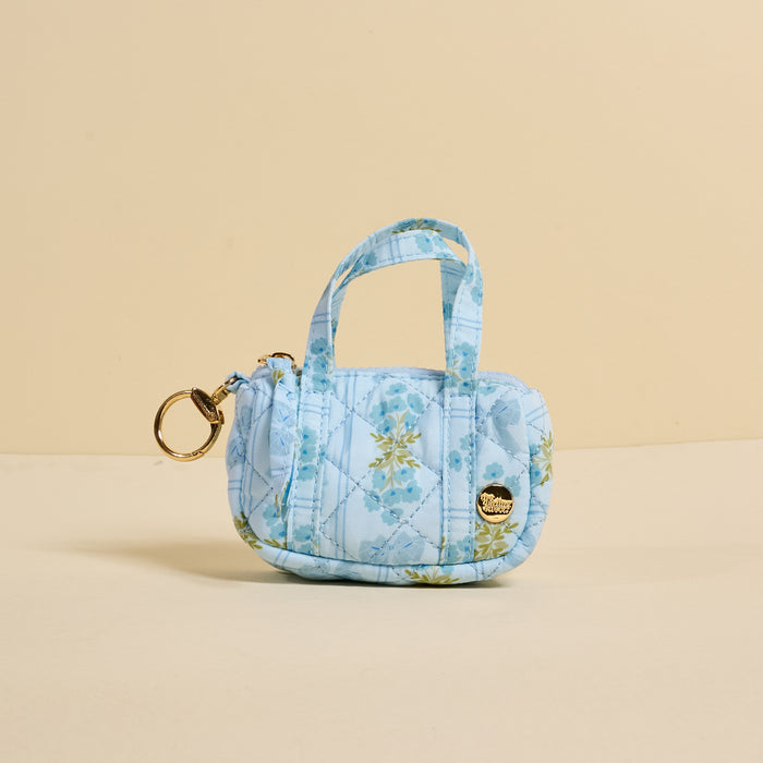 The Darling Effect's Oh So Fancy Blue Itty Bitty Duffle Bag Charm, a small quilted floral-patterned accessory with short handles, is displayed against a light yellow backdrop.