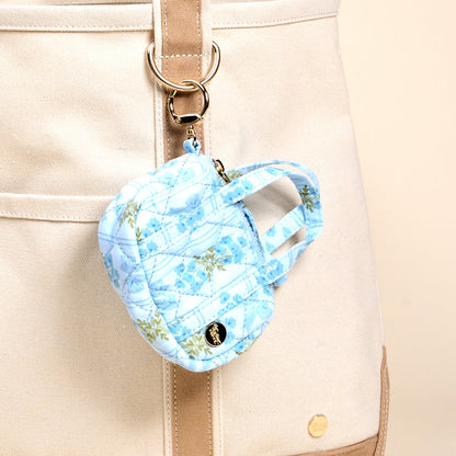 The Darling Effect’s Oh So Fancy Blue Itty Bitty Duffle Bag Charm, a quilted mini backpack keychain, is attached to a beige canvas tote with a gold keyring for the perfect stylish accessory.