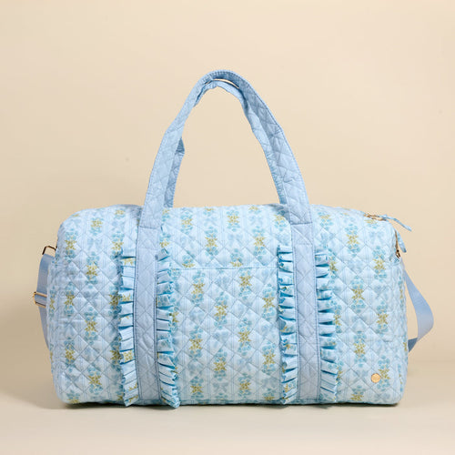 The Darling Effect Oh So Fancy Blue On-the-Go Quilted Weekender Bag features a light blue floral design, front ruffles, dual handles, a long adjustable strap, and a zippered top—spacious for travel—shown on a plain beige background.