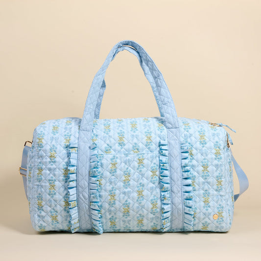 The Darling Effect Oh So Fancy Blue On-the-Go Quilted Weekender Bag features a light blue floral design, front ruffles, dual handles, a long adjustable strap, and a zippered top—spacious for travel—shown on a plain beige background.