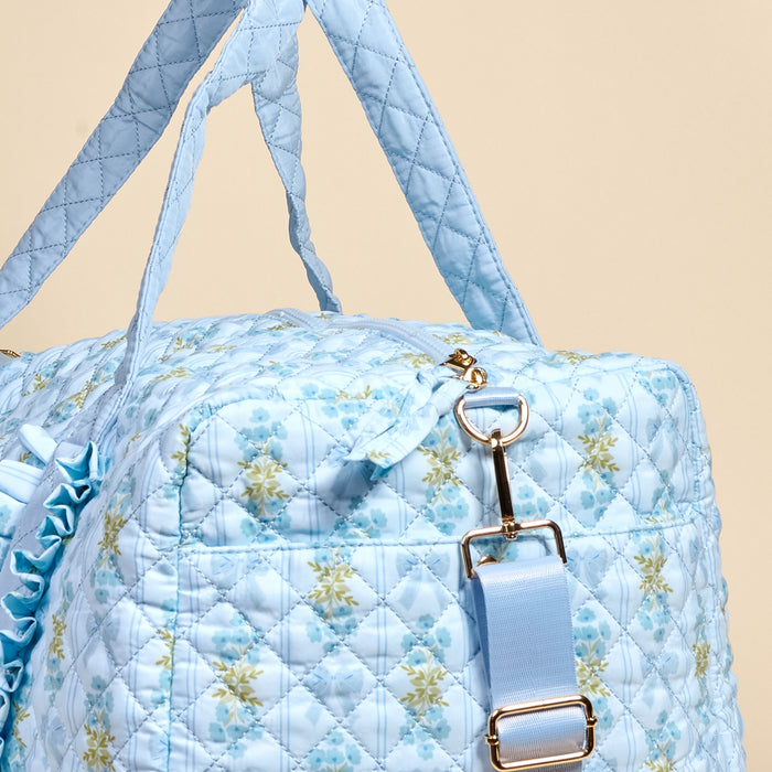 A close-up of The Darling Effect's Oh So Fancy Blue On-the-Go Quilted Weekender Bag, featuring a floral pattern, gold hardware, and matching blue straps, set against a beige background.