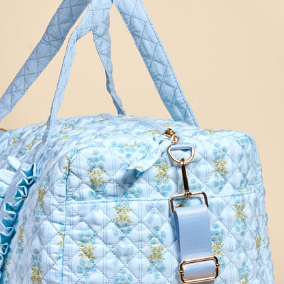 A close-up of The Darling Effect's Oh So Fancy Blue On-the-Go Quilted Weekender Bag, featuring a floral pattern, gold hardware, and matching blue straps, set against a beige background.