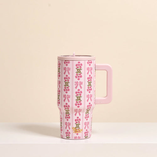 The-darling-effect’s Oh So Fancy Pink 20 oz Lifestyle Flip Straw Tumbler, featuring pink bows and rose clusters with a large handle, sits on a white surface. This insulated stainless steel tumbler keeps drinks at the perfect temperature.