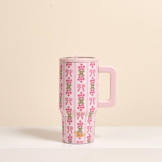 The-darling-effect’s Oh So Fancy Pink 20 oz Lifestyle Flip Straw Tumbler, featuring pink bows and rose clusters with a large handle, sits on a white surface. This insulated stainless steel tumbler keeps drinks at the perfect temperature.