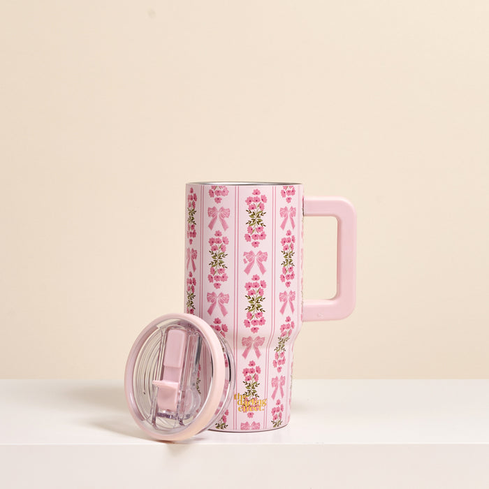 The Oh So Fancy Pink 20 oz Lifestyle Flip Straw Tumbler by the-darling-effect, featuring a pink handle, lid, bow and flower designs, stands on a white surface with its clear lid beside it against a beige background.