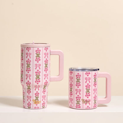 Two pink the-darling-effect Oh So Fancy Pink 20 oz Lifestyle Flip Straw Tumblers with large handles and lids rest on a white surface, both decorated with vertical stripes, pink bows, and floral patterns—the tall tumbler is on the left.