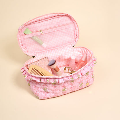The Darling Effect’s Oh So Fancy Pink Glam & Go Quilted Cosmetic Bag, featuring floral patterns and ruffled edges, is open to reveal neatly arranged makeup brushes, lip products, a comb, and a tube on a light beige background.
