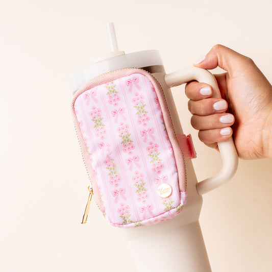 A hand holds a beige tumbler with a handle and straw, featuring the-darling-effect’s Oh So Fancy Pink Tumbler Fanny Pack—a small, water-resistant accessory with floral and bow patterns. The person’s nails are neatly manicured in light pink.