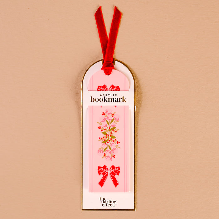 The Oh So Merry Acrylic Bookmark by The Darling Effect features a pink acrylic base with a floral design and red bows, attached to a luxurious velvet ribbon tassel. This vibrant bookmark comes on white and gold packaging labeled “Acrylic Bookmark” and “The Darling Effect.”.
