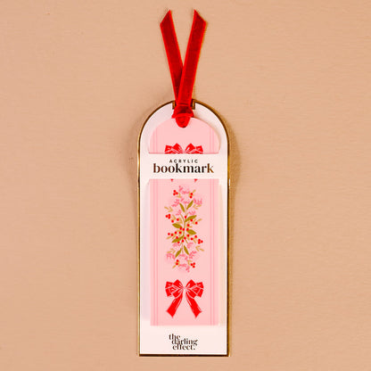 The Oh So Merry Acrylic Bookmark by The Darling Effect features a pink acrylic base with a floral design and red bows, attached to a luxurious velvet ribbon tassel. This vibrant bookmark comes on white and gold packaging labeled “Acrylic Bookmark” and “The Darling Effect.”.