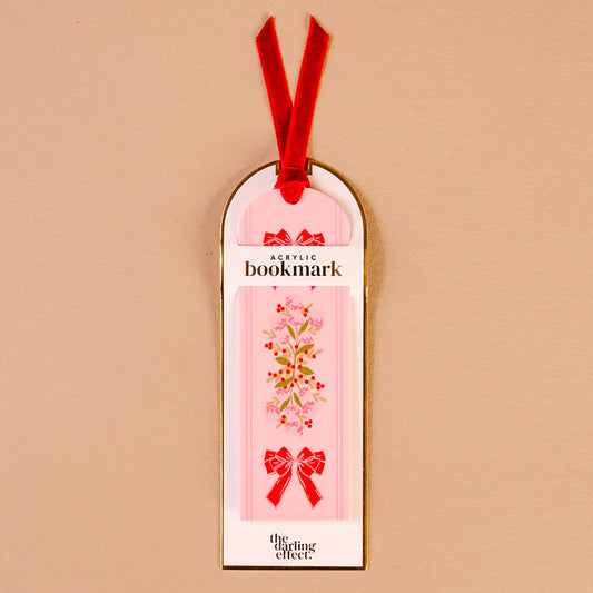 The Oh So Merry Acrylic Bookmark by The Darling Effect features a pink acrylic base with a floral design and red bows, attached to a luxurious velvet ribbon tassel. This vibrant bookmark comes on white and gold packaging labeled “Acrylic Bookmark” and “The Darling Effect.”.