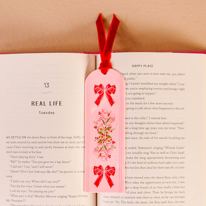 The Oh So Merry Acrylic Bookmark by The Darling Effect, featuring a vibrant floral design and a soft red velvet ribbon tassel, rests on an open book to mark the page titled REAL LIFE and HAPPY PLACE.