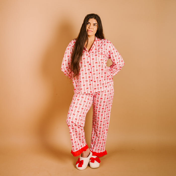 A person with long brown hair stands against a tan background wearing The Darling Effect's Oh So Merry Sweet Dreams Pajama Pants Set—pink pajamas with a red and white pattern—and white slippers with red bows.