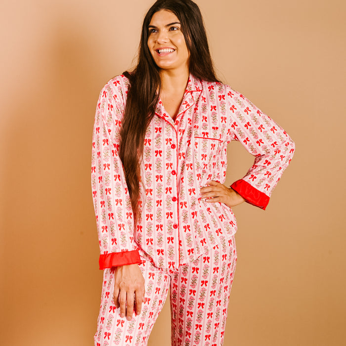 A woman with long brown hair smiles while wearing The Darling Effect's Oh So Merry Sweet Dreams Pajama Pants Set, featuring red bows and cuffs in ultra-soft fabric, standing against a beige background.